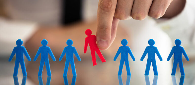 A hand picking up a small red plastic person out of a line of small blue plastic people